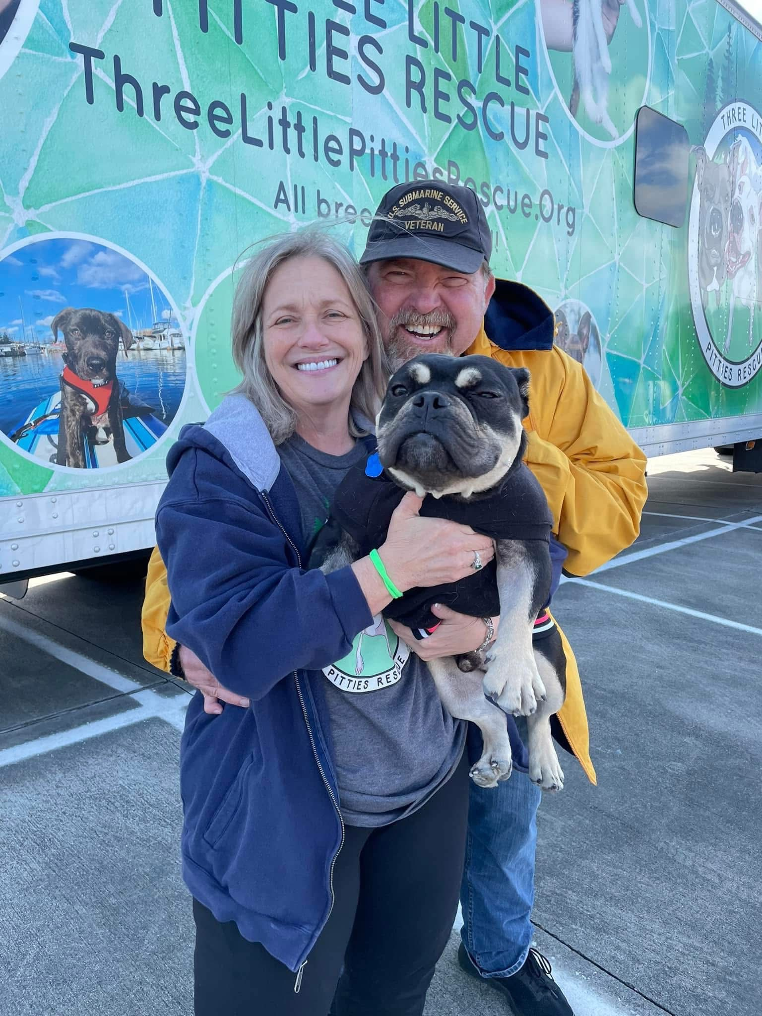 A smiling couple holding a dog beside a Three Little Pitties rescue vehicle